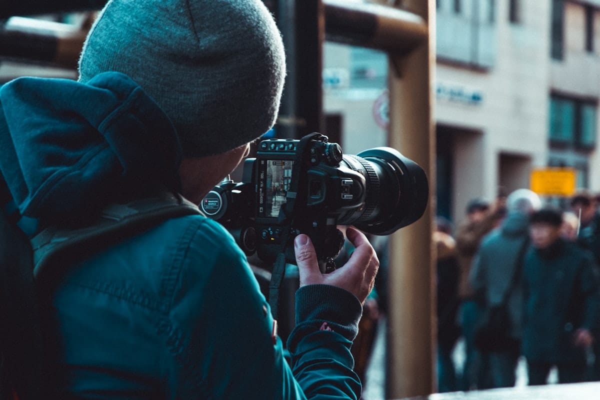 Fotograf w niebieskiej kurtce z czarnym aparatem DSLR na zewnątrz — naturalne oświetlenie dzienne, swobodna kompozycja